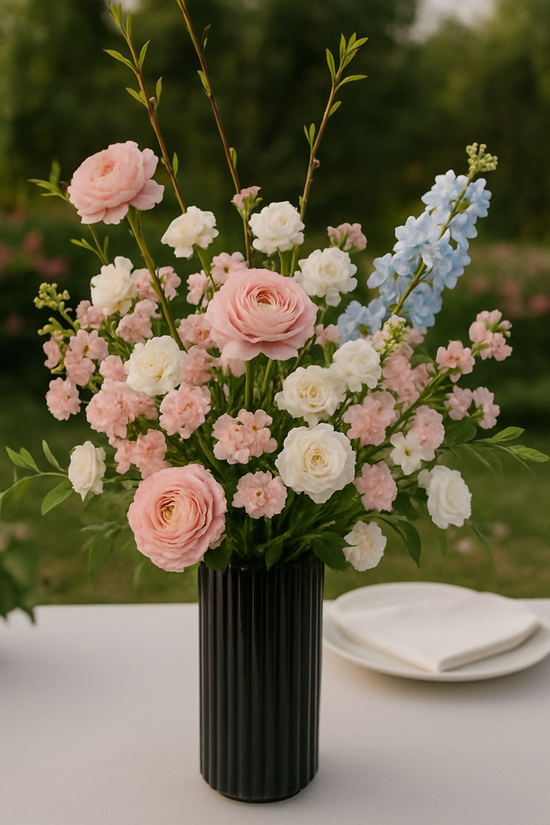 Bouquet of pink, white, and blue flowers in a black vase on a table with a blurred green background.