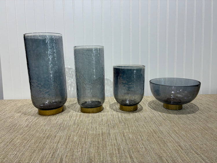 Four smoky glass vase collection of different sizes a textured surface with a white paneled wall background.