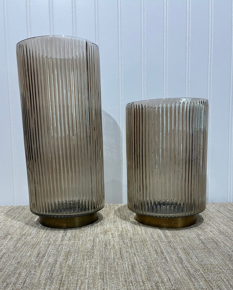Two Beige ribbed glass vases or candle holders on a textured surface with a light blue wall background.