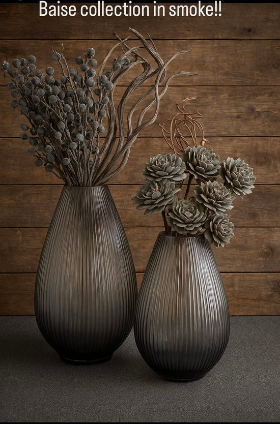 Two smoked glass vases with dried plants against a wooden wall.